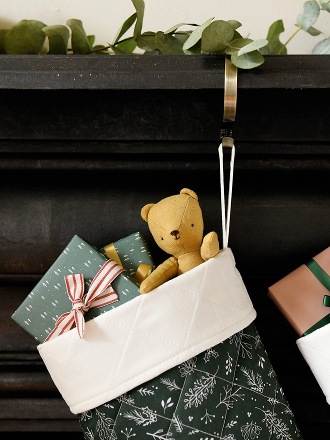 A quilted green stocking with an ivory turnover cuff hangs on a golden stocking hanger on a black mantlepiece. Small gifts and toys poke out of the top.
