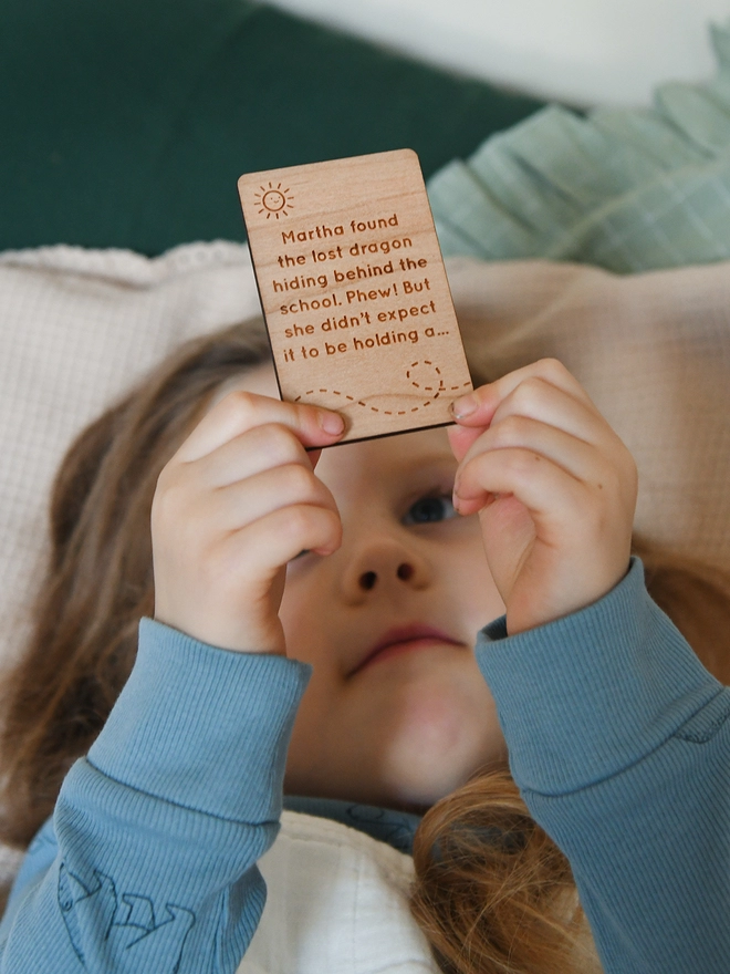 A young child wearing blue pyjamas is laying down and holding a wooden story card above her head.