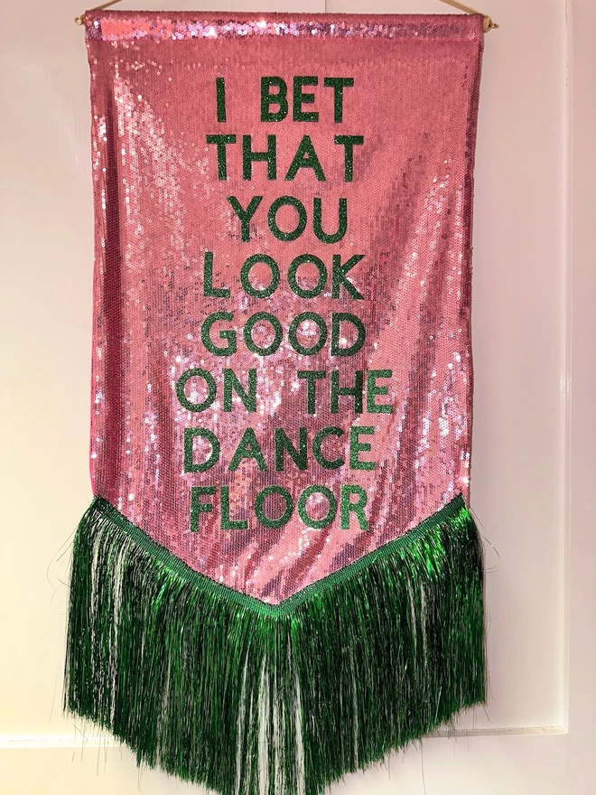 A midi customisable banner hangs from a hook. It has a light pink sequin background and a green tinsel trim along the bottom. The text is green and says 'I BET THAT YOU LOOK GOOD ON THE DANCE FLOOR'