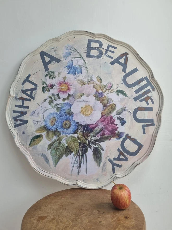 Art Panel made from a 'pie crust' table top. A pale background with mauve flowers and writing in mauve that says 'what a beautiful day'.