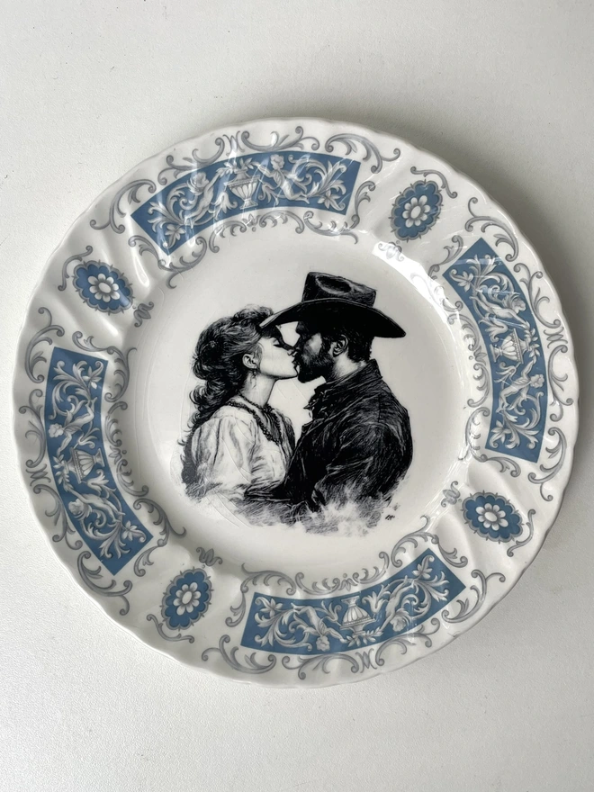 a vintage plate with an ornate border has a black and white vintage illustration of a cowboy and cowgirl sharing a kiss in the centre