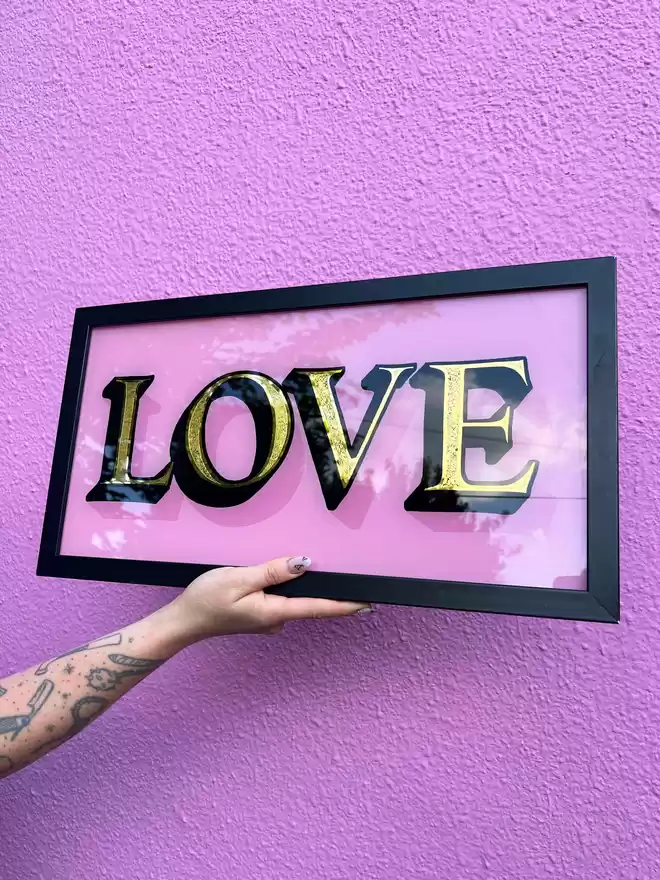 Hand-painted and gilded glass piece, spelling out LOVE. Gold leaf fill with black outlines and shade, with a baby pink background and cast shade. The piece is held up by a tattooed arm, in front of a matching pink wall.
