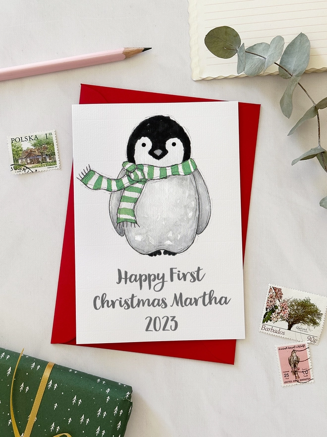 A first Christmas greetings card with an illustrated penguin design lays on a red envelope beside various stationery items on a white desk.
