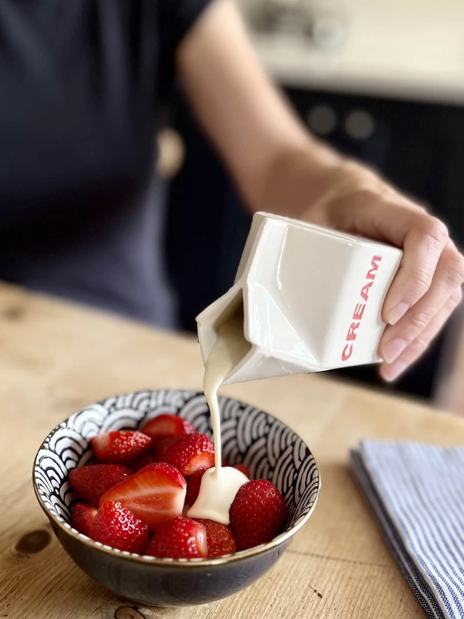 A small handmade ceramic carton is pouring cream over a bowl of fresh strawberries.