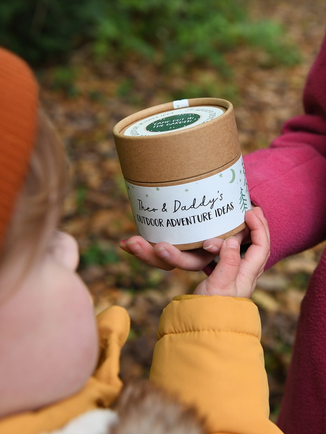 A young child wearing a yellow coat holds a cardboard jar with an outdoor adventure ideas label. Another child I na pink coat has her hand on the jar too.