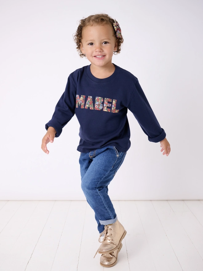 a 3 year old girl wearing a navy long sleeve t-shirt with her name Mabel cut from Liberty floral fabric and sewn onto the front 