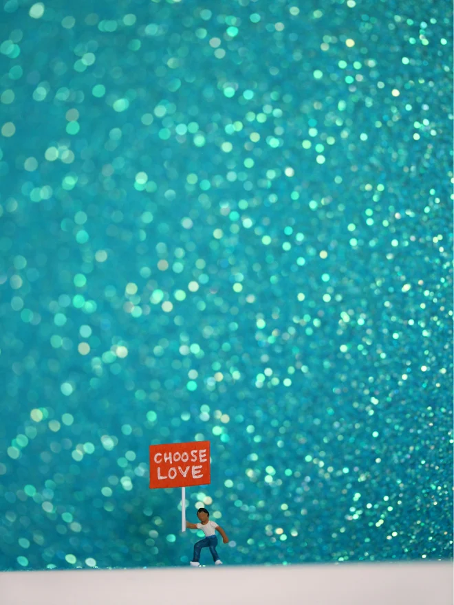 Miniature scene in an artbox showing a tiny child is holding a protest sign saying "Choose Love" against a sparkling blue backdrop. (close up)