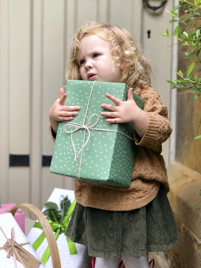A toddler is holding a gift wrapped in green wrapping paper with a tiny tree design.