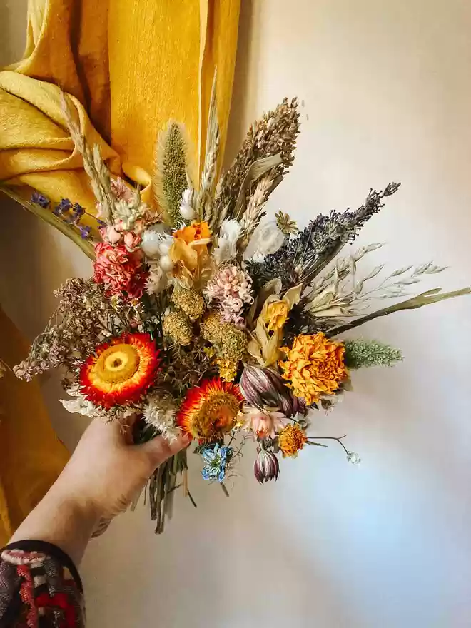 Natural, Seasonal Dried Flower Bouquet seen held by a hand with a mustard coloured fabric behind.