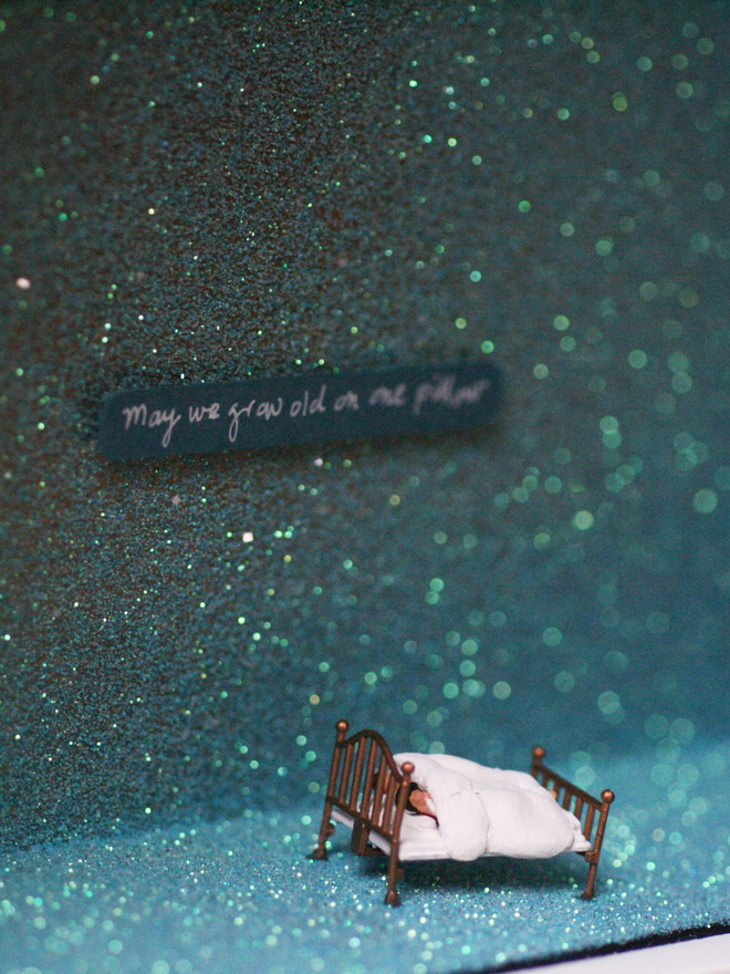 Miniature scene in an artbox showing a tiny man and woman lying in bed against a sparkling backdrop evoking the night sky. The words “May we grow old on one pillow” are written on the back wall.