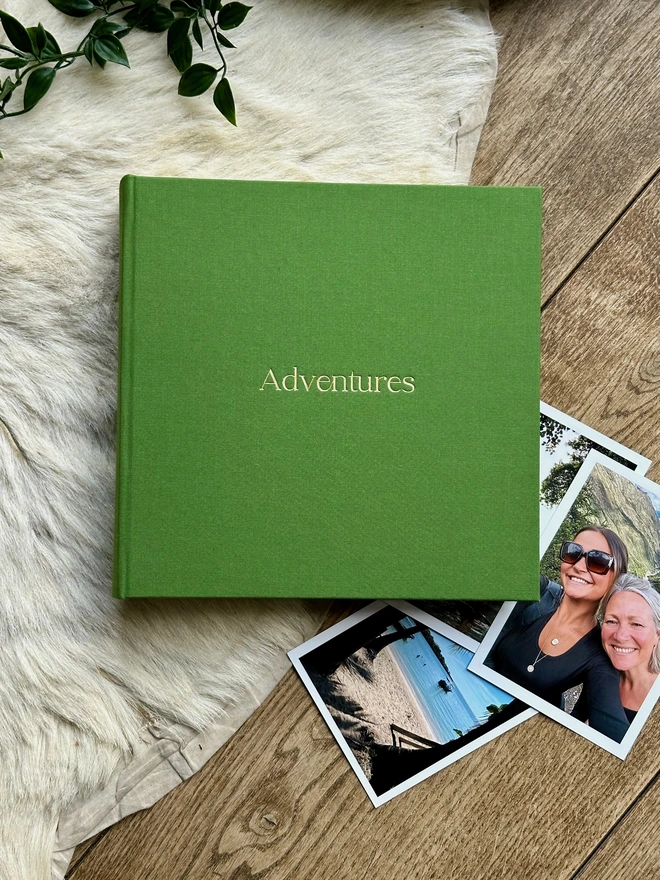 A hand-bound green photo album printed with the words 'Adventures' in gold lettering rests on a bookshelf next to rows of red books.