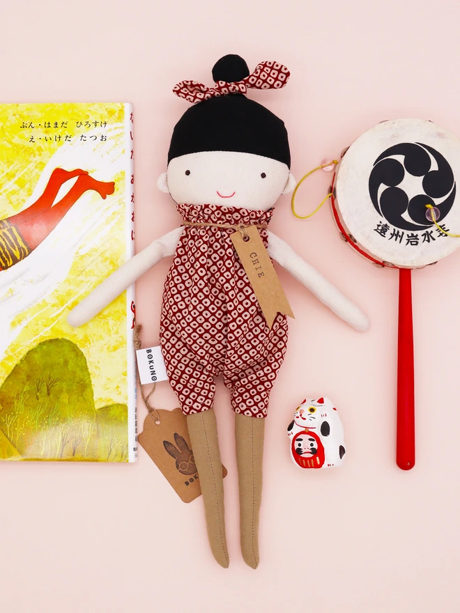 A handmade doll wearing a red and white Japanese shibori patterned jumpsuit with a matching bow in her black hair. The doll is surrounded by Japanese children's book, maneki-neko cat and drum.