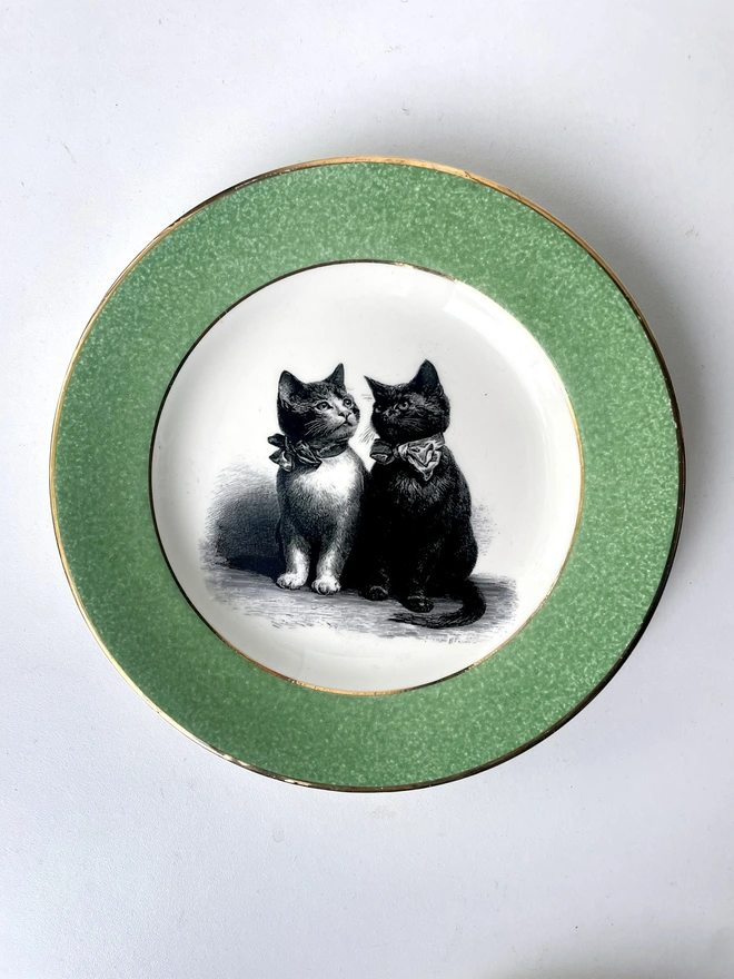 a vintage plate with a pea green border with gold trim and in the centre is an illustration of a pair of Victorian kittens