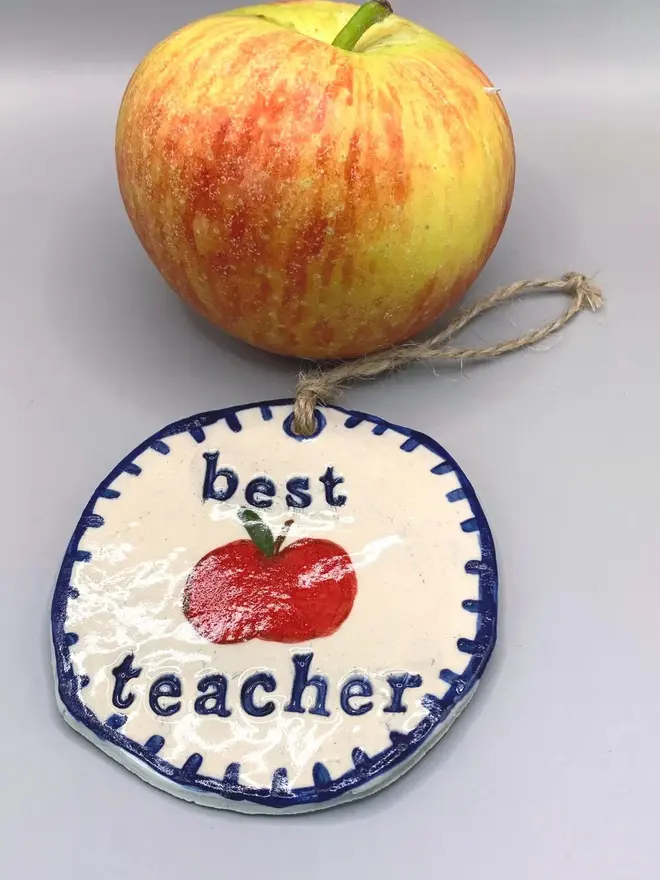 'Best Teacher' Handcrafted Ceramic Ornament, a ceramic apple ornament sitting on a plain white surface joined by an apple. 