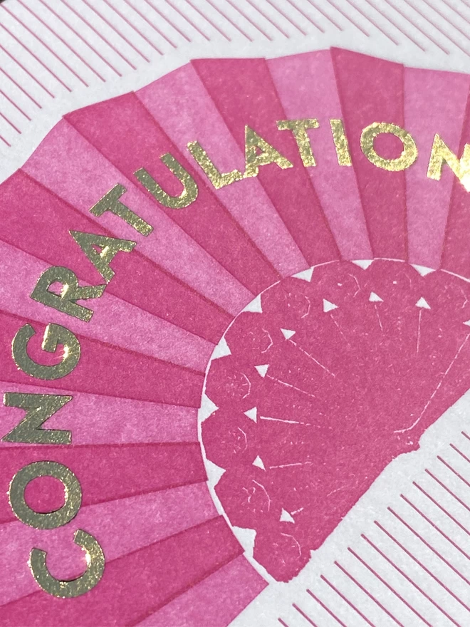 Close up of a pink and white striped card with a pink fan on it and gold text reading 'Congratulations'.