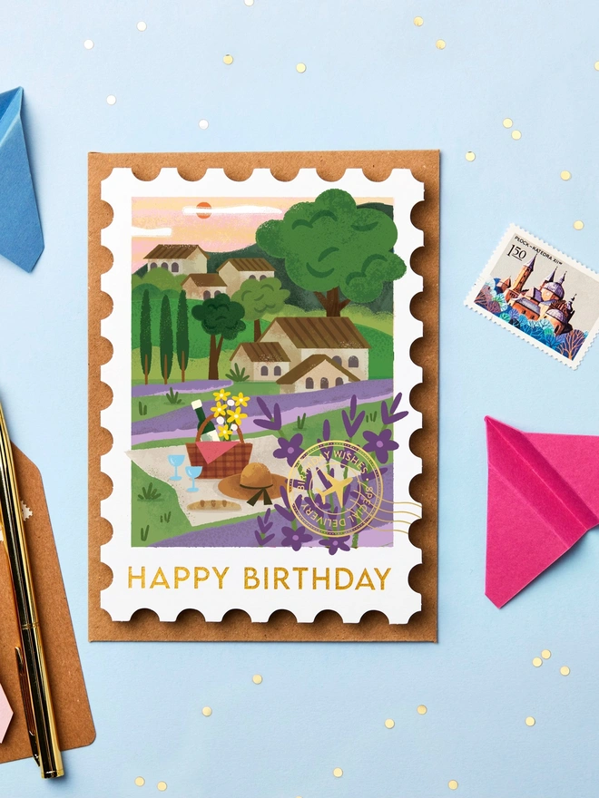 Stamp-shaped greeting card featuring an illustration of Provence. The card is on a blue background with paper aeroplanes and stationery around it.