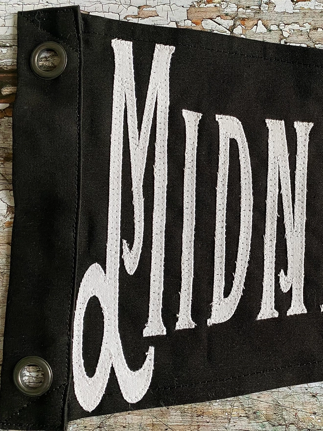 A close up of a section of black Midnight Margarita pennant flag showing the letters in white MIDN