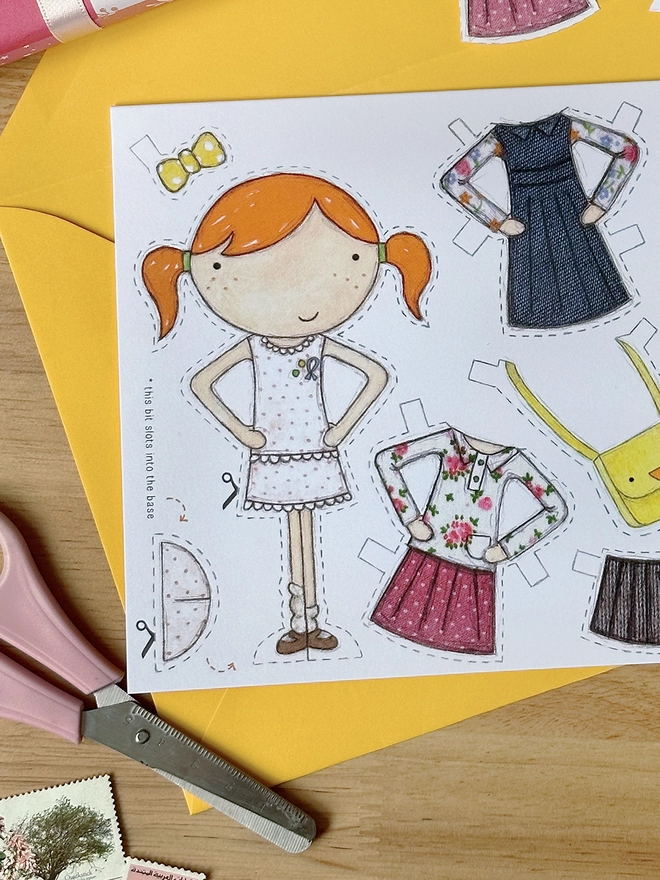 A paper doll greetings card with a paper doll and several outfits on the card lays on a yellow envelope on a wooden desk.