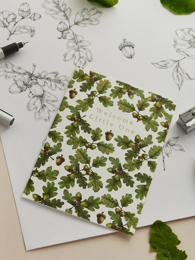 'welcome little one' illustrated oak leaf card. a white card illustrated with oak leaves photographed with a sheet of line drawings.