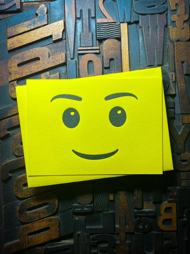A bright yellow letterpress card with a smiling face emoji is placed on top of a matching  yellow envelope. The background consists of various wooden printing letters in different orientations and sizes.