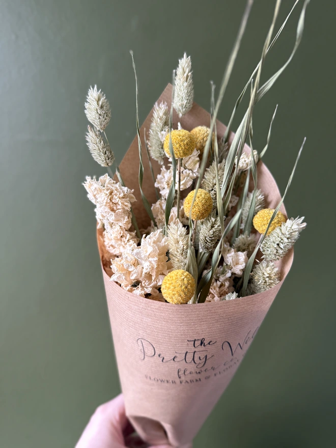 a bunch of dried flowers in bright colours wrapped in black tissue and kraft paper held by a person in a mustard coloured jumper with their left arm against a green wall