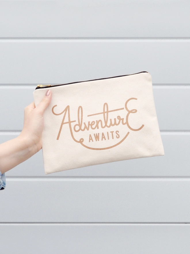 a hand holding out a natural canvas zip pouch with the words adventure awaits