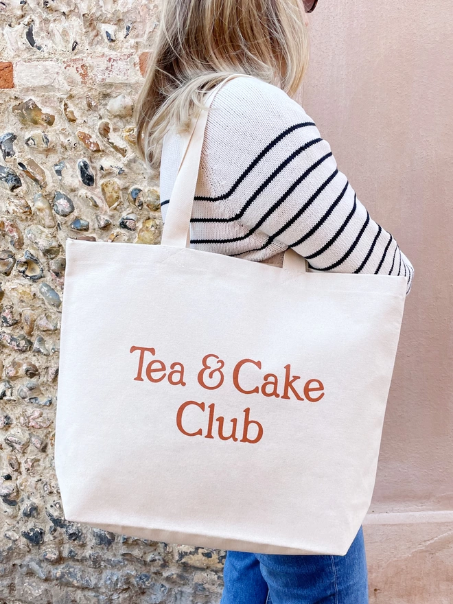 model holding a large natural canvas tote bag bearing the words tea and cake club