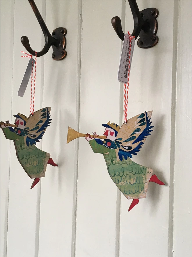 Two Joyful Angel illustrated wooden Christmas decorations in green, gold and red. The decorations hang on vintage hooks against a green panelled wall. aganst a white background.