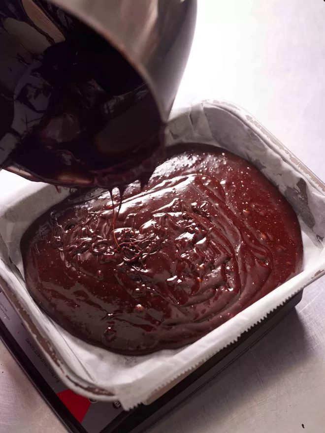 Pouring gluten free classic fudge brownie mixture into a baking tray to measure in the bakery