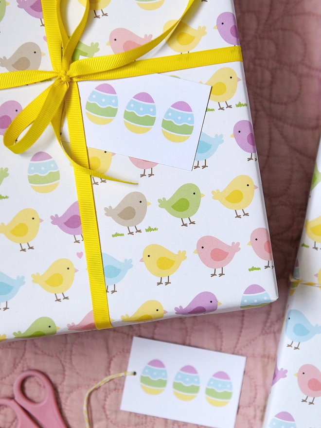 A gift wrapped in pastel spring chicks wrapping paper tied with yellow ribbon is on a pink quilt.