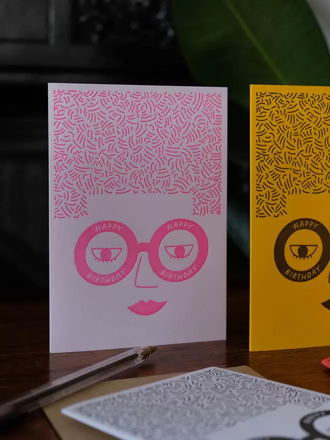 Pink card with a pink ladies face wearing glasses saying ' Happy Birthday' and big square hair. 