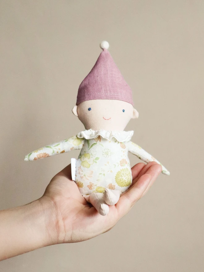 A handmade Spring Elf Doll with light skin, a soft pink floral print body, and a lavender-pink hat topped with a white pompom. The doll has a gentle smile and wears a ruffled white collar.