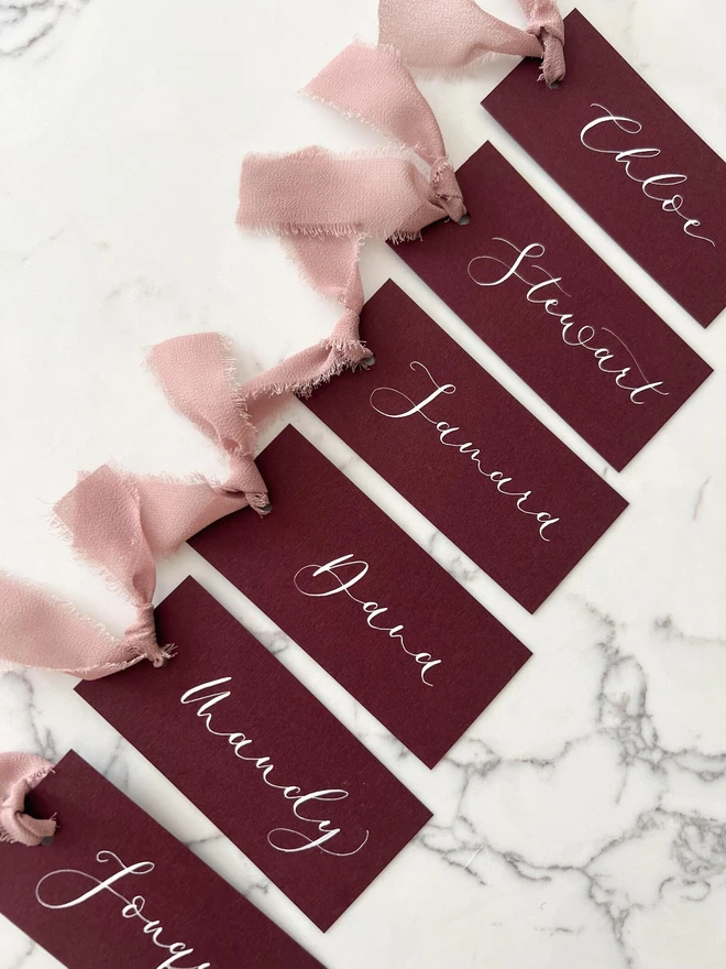 Personalised Calligraphy Wedding Burgundy Place Cards, burgundy place cards set together on a wooden table with chiffon plum ribbons. 