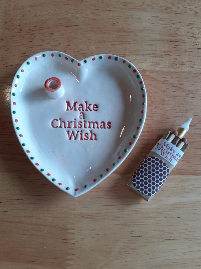 Handmade white ceramic heart-shaped Christmas Wish Dish with red lettering and alternating red and green dots, displayed beside a small box containing a gold candle and matches.