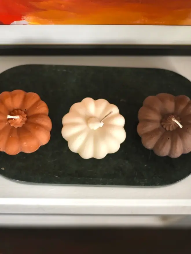 Medium Rust Wax Pumpkin Candle. Three pumpkin candles in terracotta, cream and chocolate brown lined up on a green marble tray beneath a colourful painting. The trio makes an elegant autumn arrangement for a shelf, sideboard or windowsill.