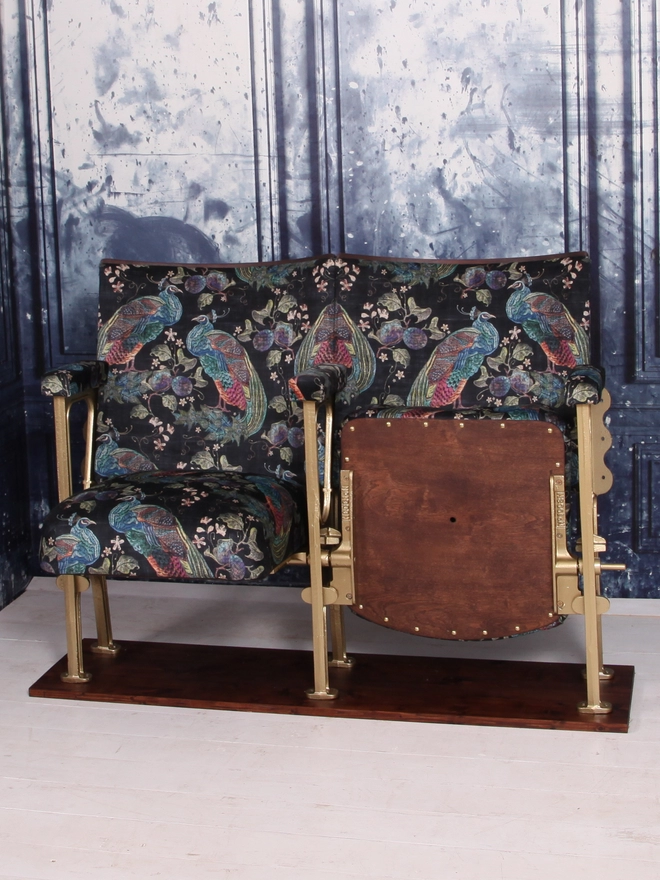Set of two vintage cinema seats against a blue marbled wall.  One seat is up and one seat is down, both seats are upholstered in a black velvet with colourful peacocks on it