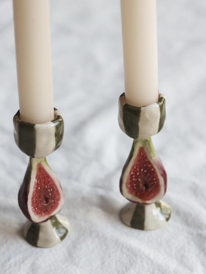 Candlesticks, a pair of candle sticks on a white tablecloth. They have small emblems of fruits or vegetables on them. 