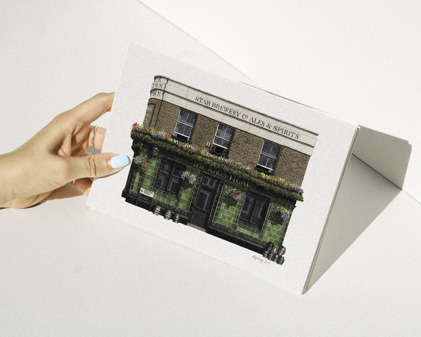A hand with blue nail polish holding a print featuring an illustration of The Pelton Arms pub against a white background.