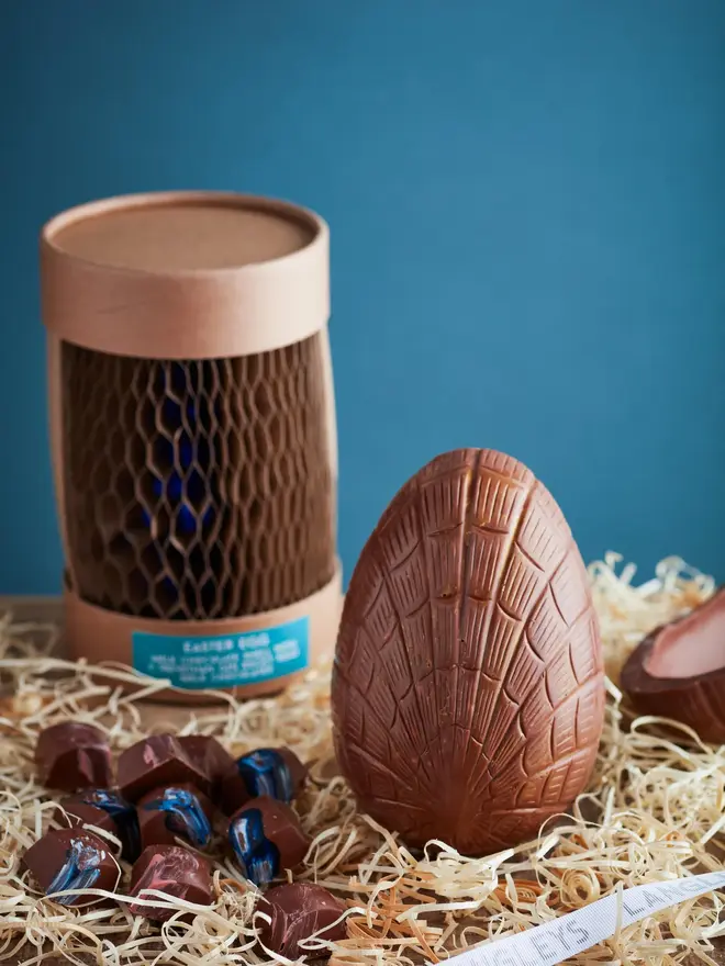 Extra Thick Milk Chocolate Easter Egg With Trevethan Gin, a chocolate egg sitting on a hay surface against a blue backdrop. It is accompanied by the packaging and some small chocolates from within. 
