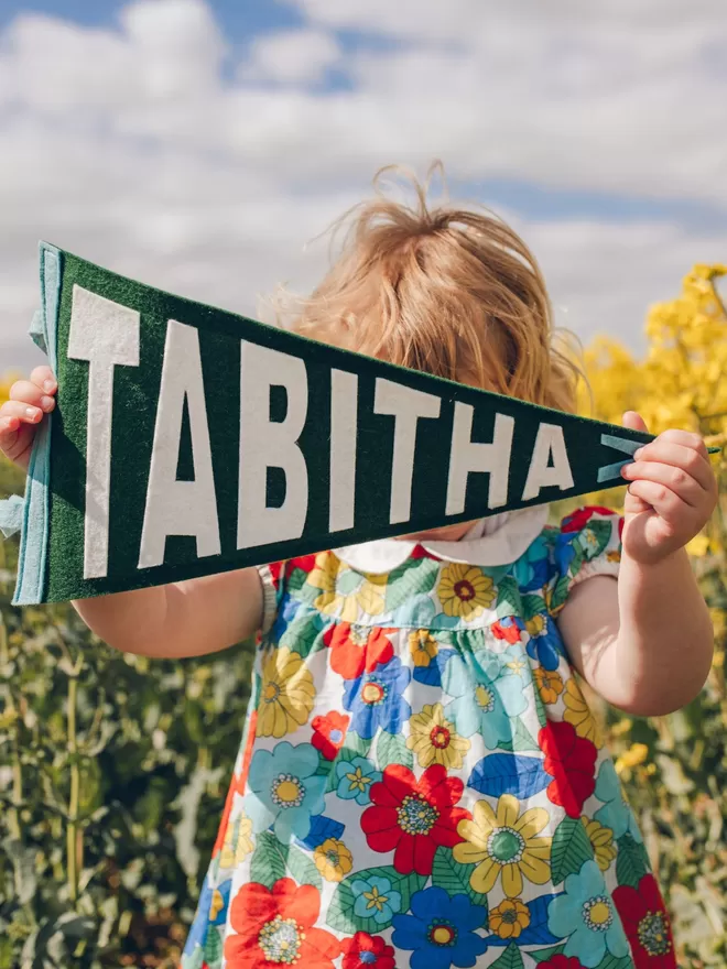 A young girl in a flowery dress holding up a pennant flag with the name Tabitha.