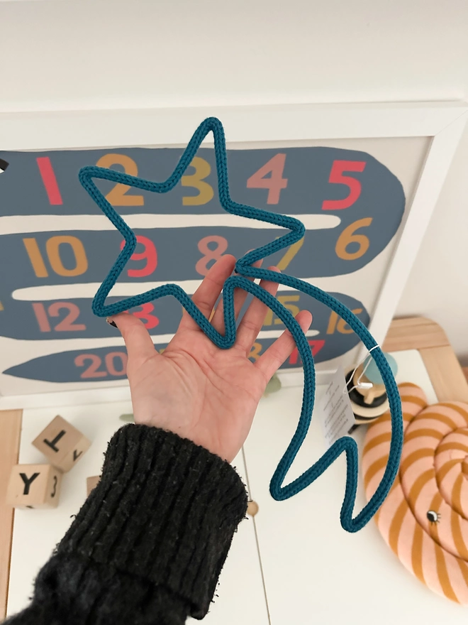 A hand holding a shooting star made from knitted cord and wire over a table with children's toys, table and wall art. 
