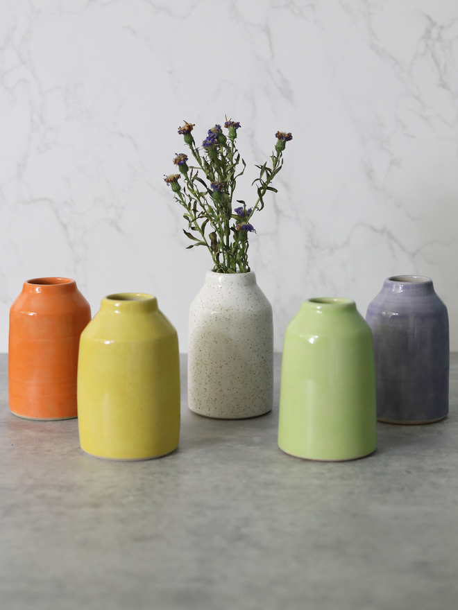 Five handmade ceramic bud vases arranged in a row on a grey surface, showing the full colour range of orange, yellow, speckled white, spring green, and lavender, with dried purple wildflowers in the central white vase, against a white marble background