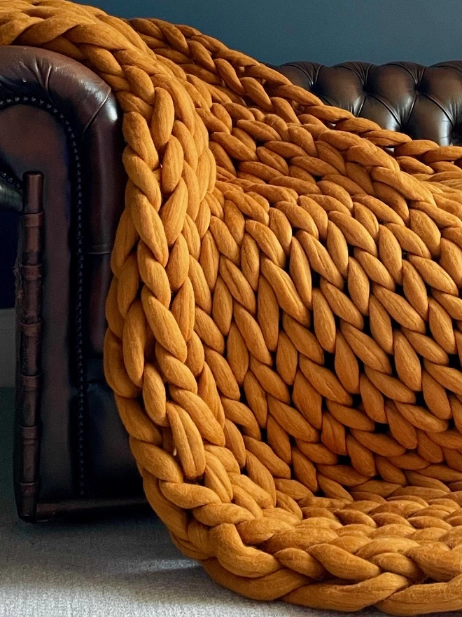 A close up of A rainbow merino arm knitted blanket cascading down the side of a brown leather Chesterfield sofa, before a dark blue wall