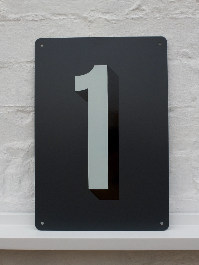 Pale grey and black house number 1, on anthracite grey metal plaque, against a white brick wall.