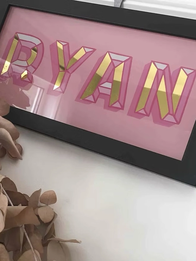 Personalised Name Wall Plaques, a pink personalised gold leaf sign sitting in a black frame hanging in front of a white wall. 