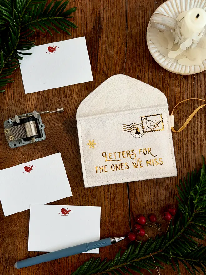 A handmade ivory felt envelope decoration with the words 'Letters for the ones we miss' in gold lays on a wooden table. Several robin notecards lay beside it.
