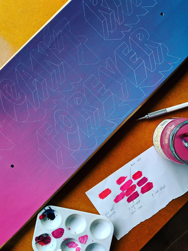 Detail of a painted skateboard showing a spray painted pink and blue background and 3d letters sketched on. An artist’s paint palette, water pot and colour tests lie alongside