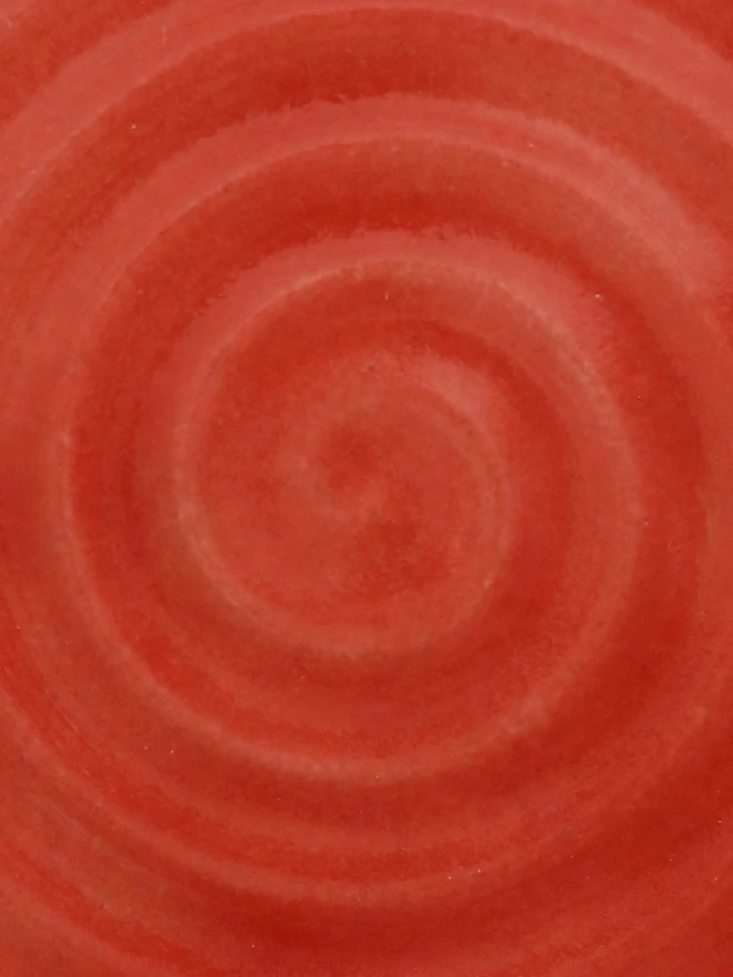 A close-up of a handmade ceramic glaze swatch in cherry red, showing a smooth concentric swirl texture with a delicate, even tone throughout