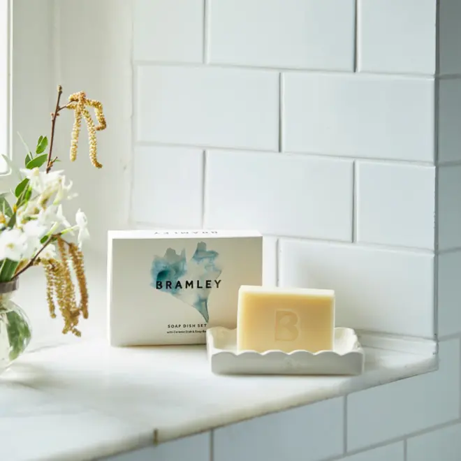 white scalloped soap dish, with off-white bar of soap, engraved with "B" logo. next to a white cardboard box with 'bramley' both sitting in white-tiled window sill