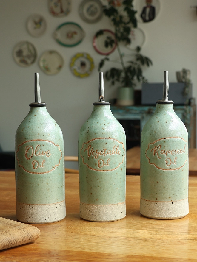 Three handmade stoneware oil pourers sat on a wooden table in a kitchen scene. The oil pourers feature the  words 'Vegetable Oil', 'Rapeseed Oil' and 'Olive Oil' and are glazed in a mint green glaze